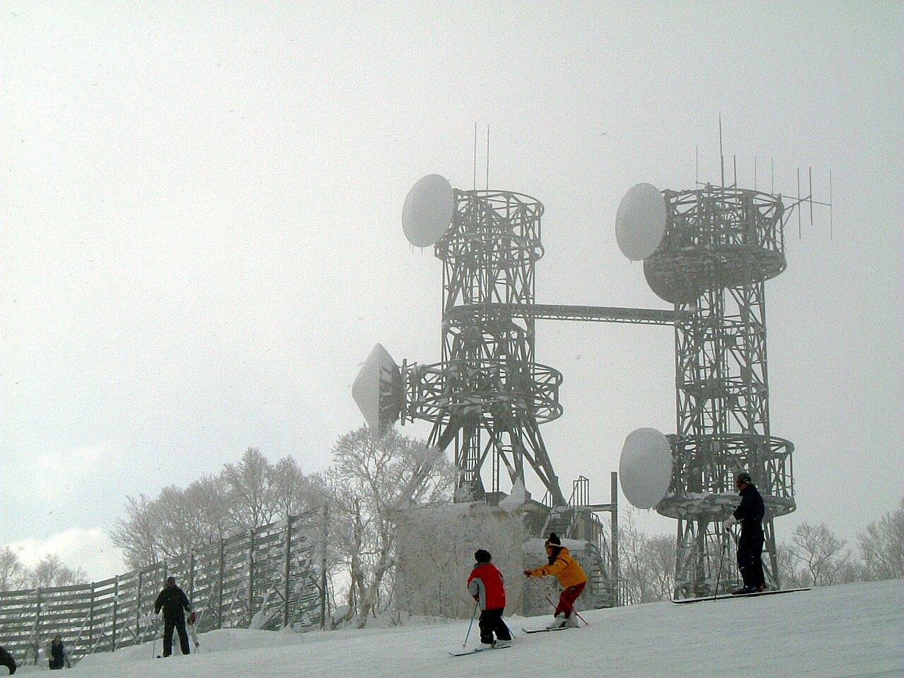 นักสกีกำลังเล่นบนลาน Sapporo Teine เห็นเสาส่งสัญญาณเคลือบหิมะบนยอดเขาเป็นฉากหลัง