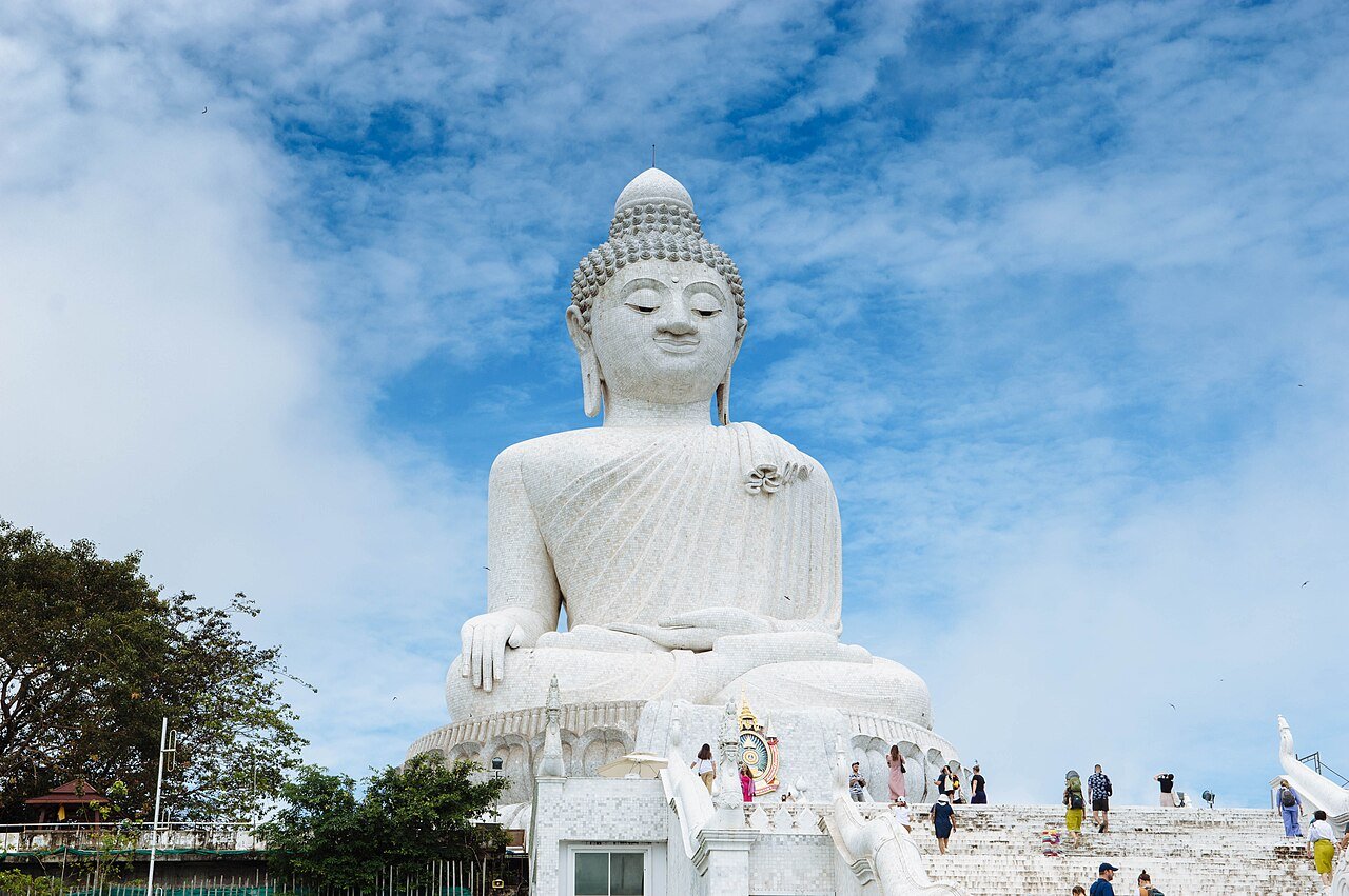 พระใหญ่ภูเก็ต — พระพุทธรูปหินอ่อนสีขาวขนาดใหญ่ประทับนั่งบนยอดเขา ท้องฟ้าสีฟ้าเมฆขาว มีนักท่องเที่ยวเดินขึ้นบันได