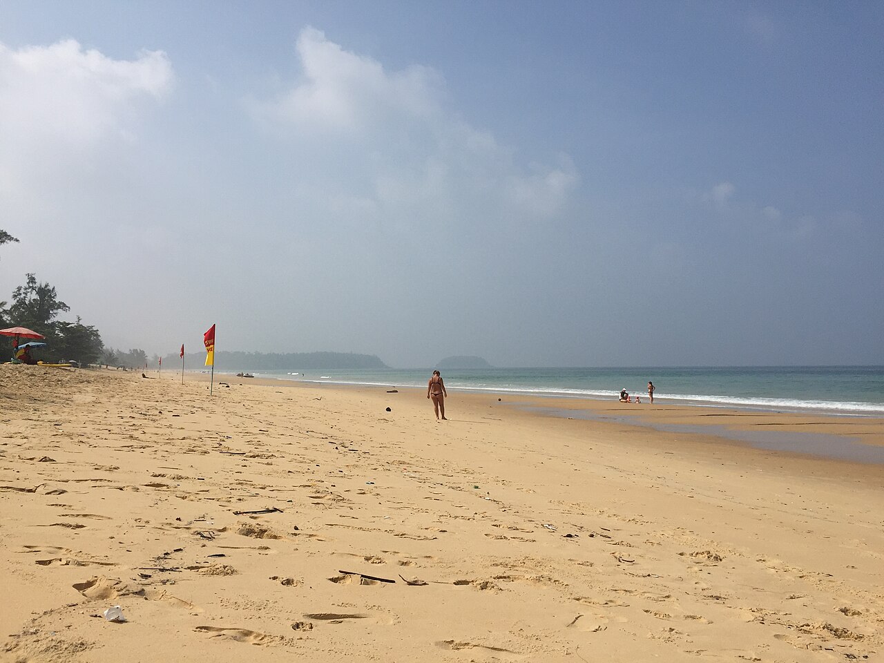 หาดกะรน ภูเก็ต — หาดทรายขาวยาวกว้างเปิดโล่ง มีธงแดงปักเตือนกระแสน้ำบนหาด ท้องฟ้าฟ้าใส