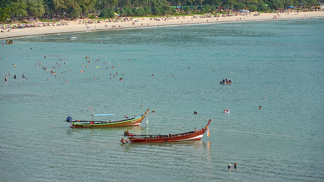 หาดกะตะ ภูเก็ต — น้ำทะเลสีเขียวมรกตใส เรือหางยาวจอดกลางน้ำ คนเล่นน้ำ และแนวหาดทรายขาวด้านหลัง