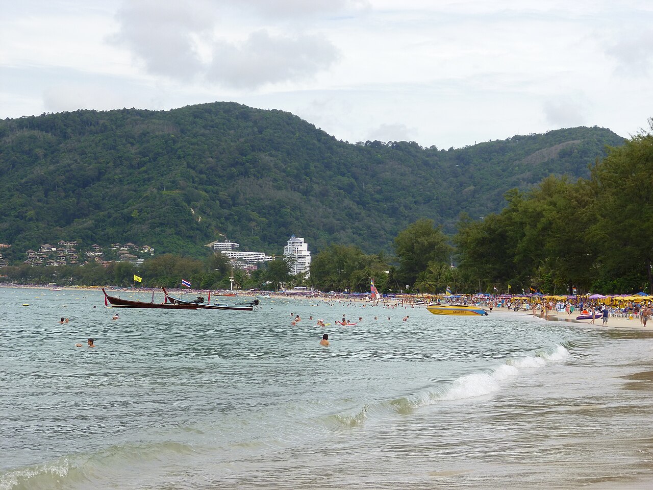 หาดป่าตอง ภูเก็ต — หาดทรายโค้งคนเล่นน้ำคึกคัก เรือหางยาวจอดริมฝั่ง ภูเขาเขียวและตึกโรงแรมเป็นฉากหลัง