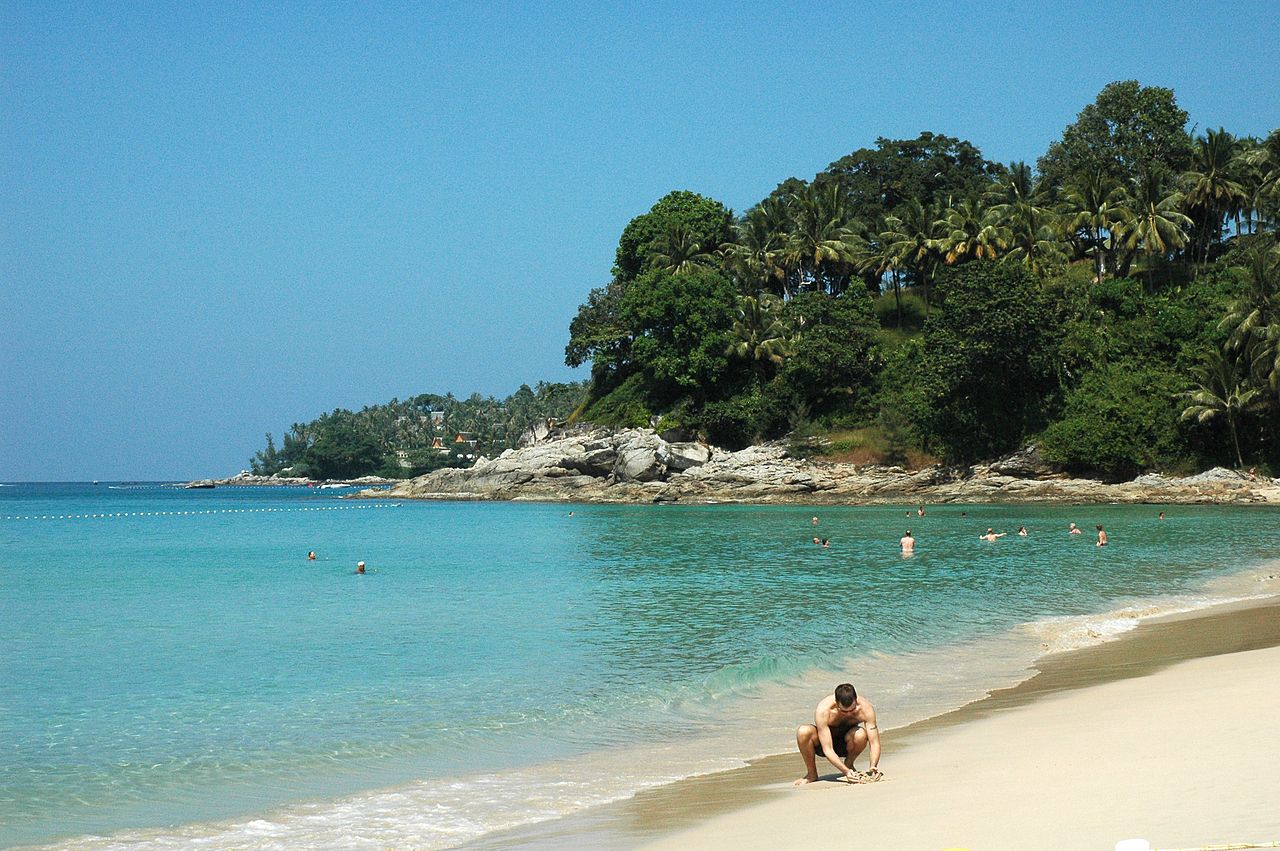 หาดสุรินทร์ ภูเก็ต — น้ำทะเลสีฟ้าครามใส หาดทรายขาว หัวหาดหินและแนวต้นมะพร้าวเขียวชอุ่ม คนเล่นน้ำหลังแนวทุ่น