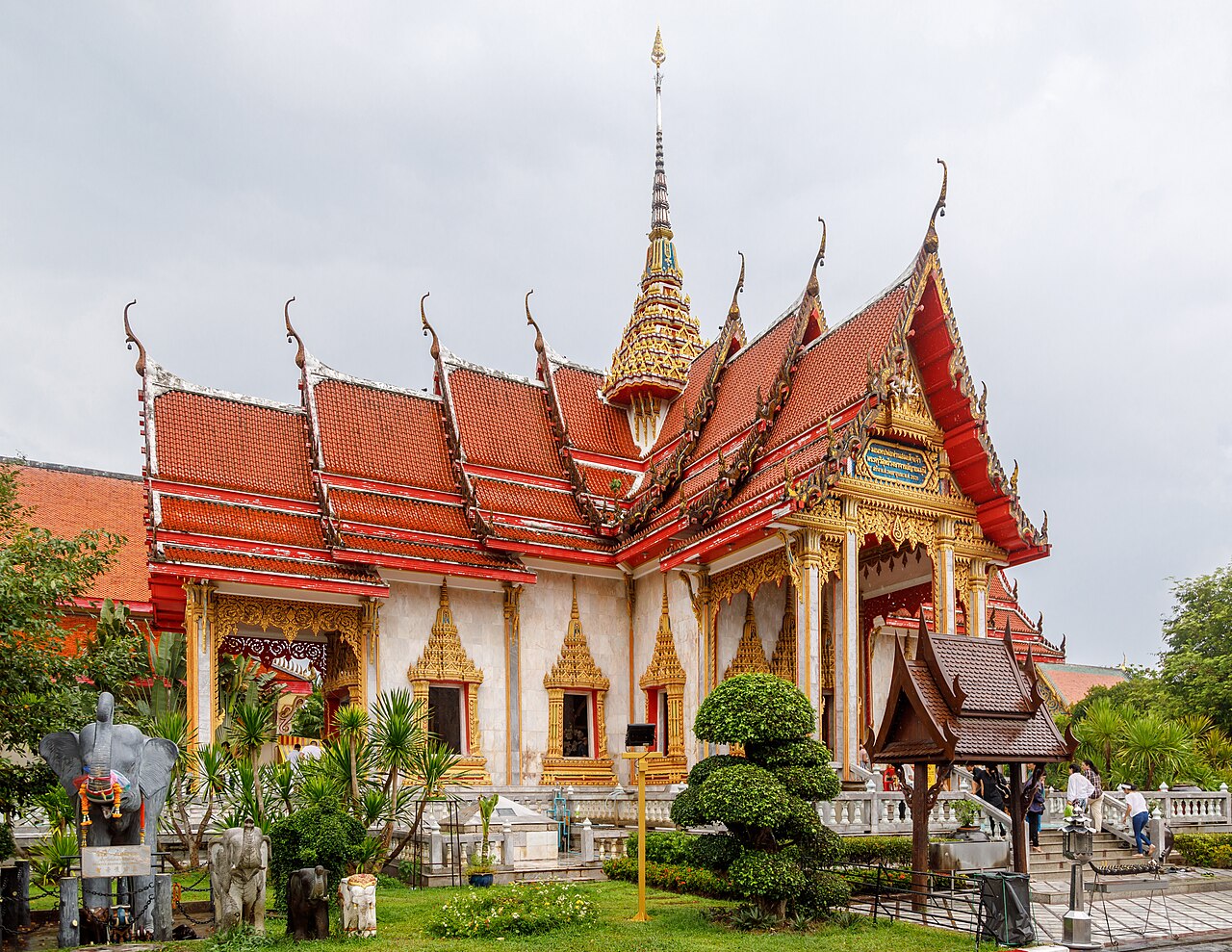วัดฉลอง ภูเก็ต — อุโบสถทรงไทยหลังคาซ้อนชั้นสีแดงทอง มีช่อฟ้าปลายแหลม ล้อมด้วยสวนเขียวและรูปปั้นช้าง
