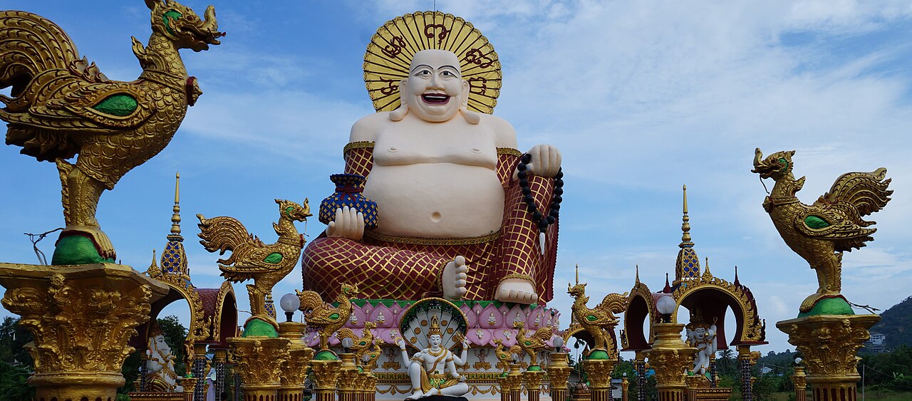 พระสังกัจจายน์องค์ยิ้มสีขาวขนาดยักษ์ที่วัดปลายแหลม เกาะสมุย ล้อมด้วยซุ้มและเสาสีทองริมบึงน้ำ