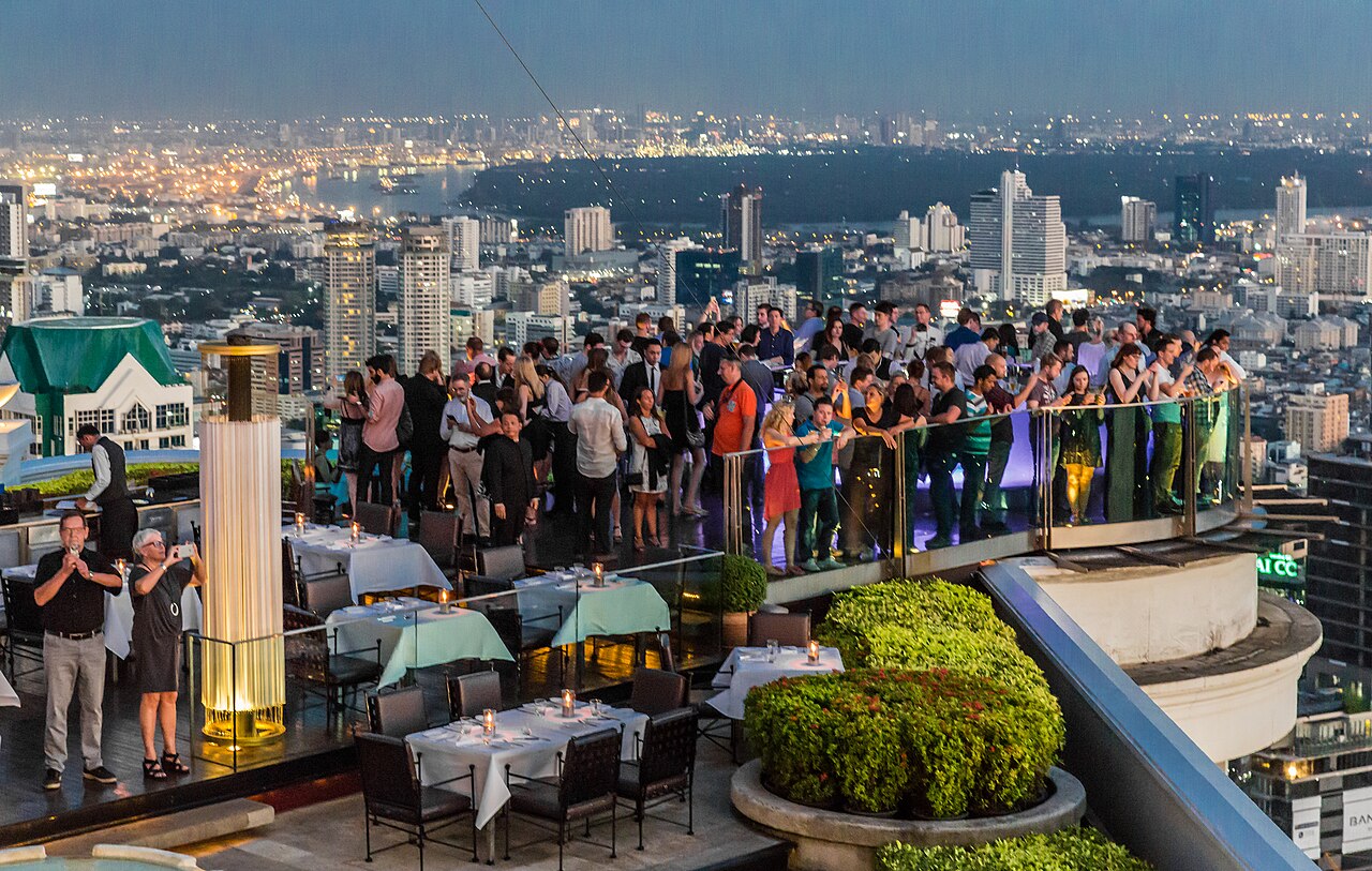 ลานดาดฟ้า Sky Bar ที่ lebua State Tower กรุงเทพ — คนยืนชมวิวแม่น้ำเจ้าพระยายามค่ำริมราวระเบียงชั้น 63 โต๊ะดินเนอร์ของห้องอาหาร Sirocco และเสาไฟสีทอง