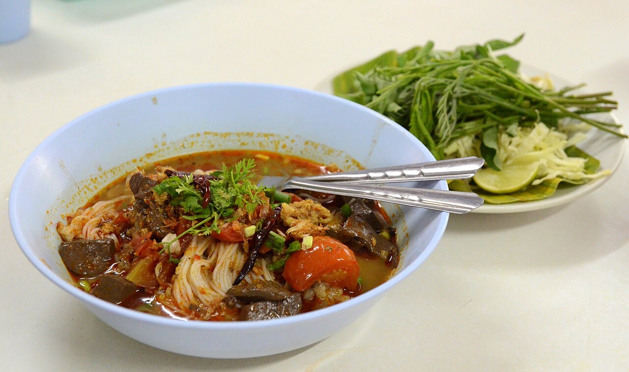 ขนมจีนน้ำเงี้ยว เส้นขนมจีนราดน้ำเงี้ยวสีแดงส้มมีมะเขือเทศและเลือด ในชาม พร้อมผักเคียงจานข้าง