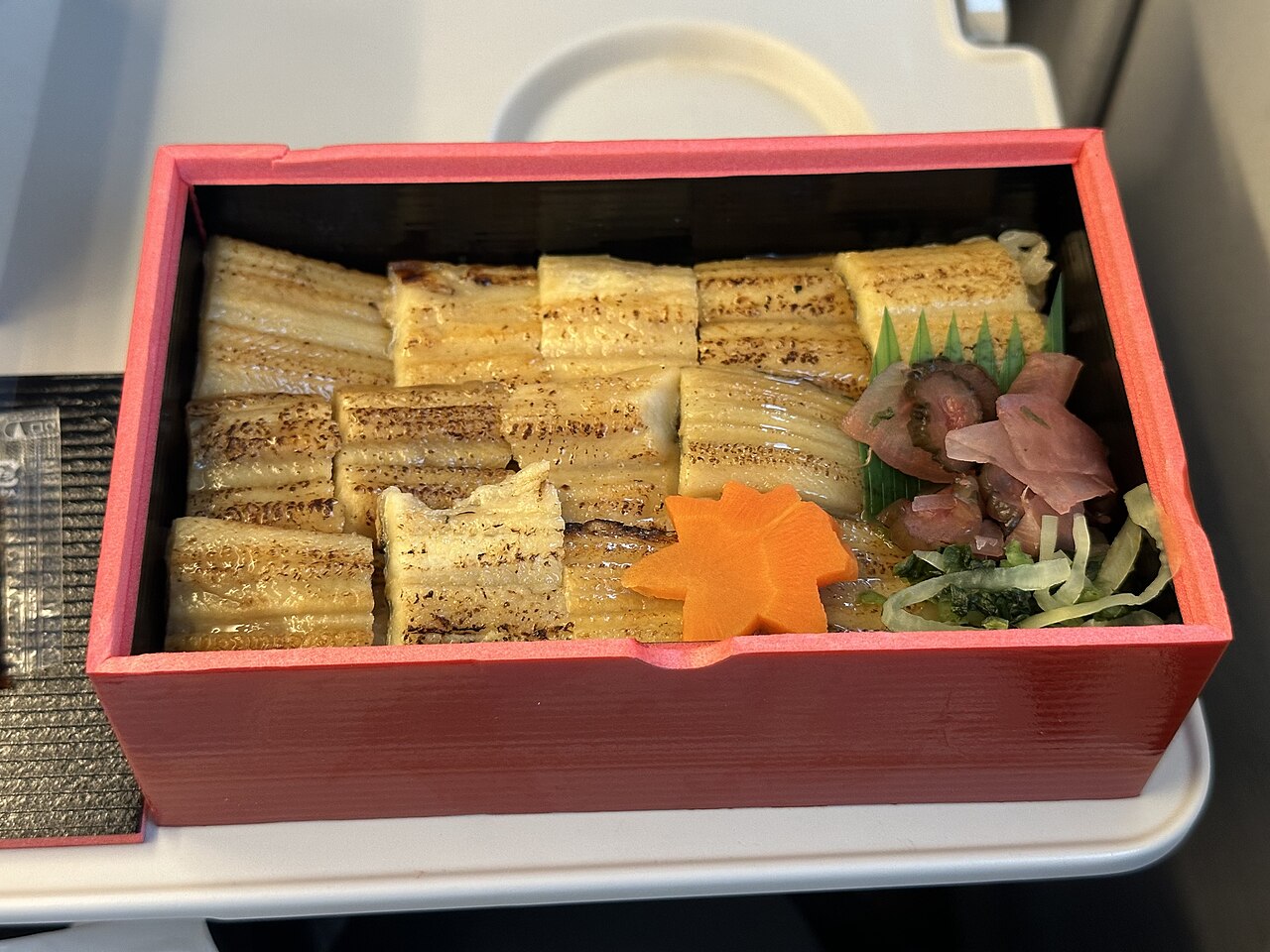 กล่องอานาโกะเมชิ — ข้าวหน้าปลาไหลทะเลย่างในกล่องเบนโต้สีแดง ชิ้นปลาไหลสีทองเรียงบนข้าว ตกแต่งด้วยดอกแครอทและใบเตยสีเขียว