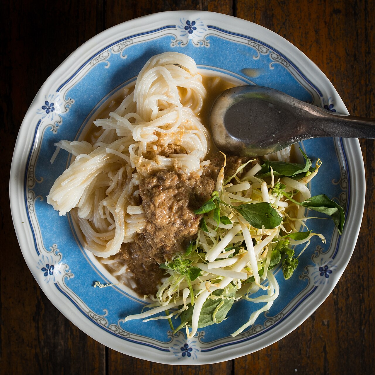 ขนมจีนราดน้ำยาเข้มข้นในจานลายคราม พร้อมถั่วงอก ใบโหระพา และผักเหนาะ