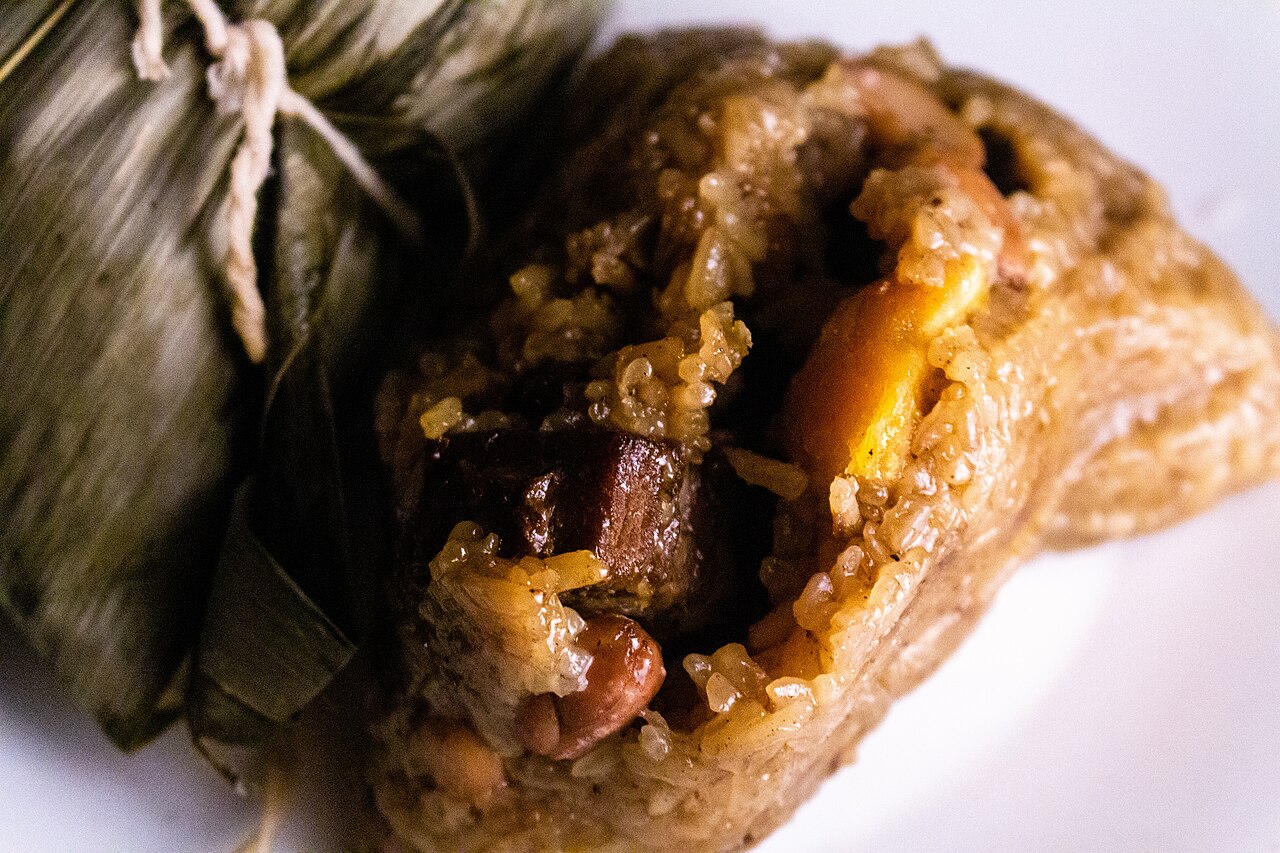 A zongzi opened to show brown sticky rice, a large chunk of braised pork belly, peanuts and an orange salted egg yolk, wrapped in a bamboo leaf
