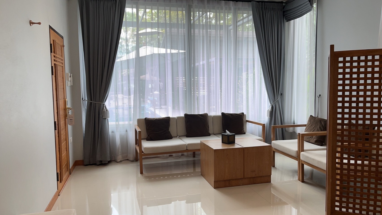 มุมนั่งเล่นในห้องพัก Look at Home Lamphun โซฟาไม้สัก ผ้าม่านเทา กระจกบานสูงมองออกสวน