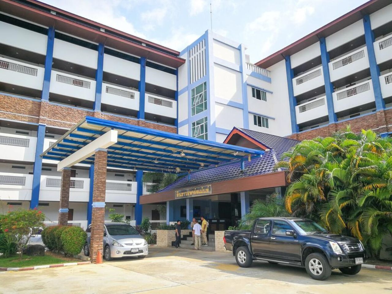 โถงทางเข้า Din Khao Apartment นราธิวาส มีหลังคาคลุมรถยื่นออกมา ป้ายชื่อโรงแรมอยู่เหนือประตูทางเข้า ตัวตึกขาวคาดแถบน้ำเงิน มีรถจอดหน้าตึก