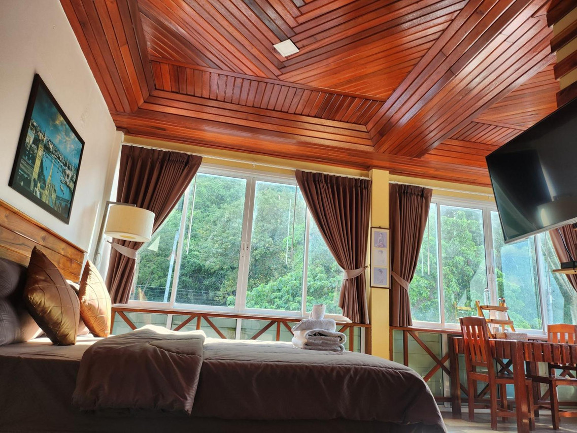 ห้องวิวเขาอีกมุมของ Mountain View Guest House พัทลุง เพดานไม้สัก เสาลายไม้ ผนังขาว โคมไฟหัวเตียง หน้าต่างกระจกยาวมองเห็นป่าเขา มีโต๊ะกินข้าวริมหน้าต่าง
