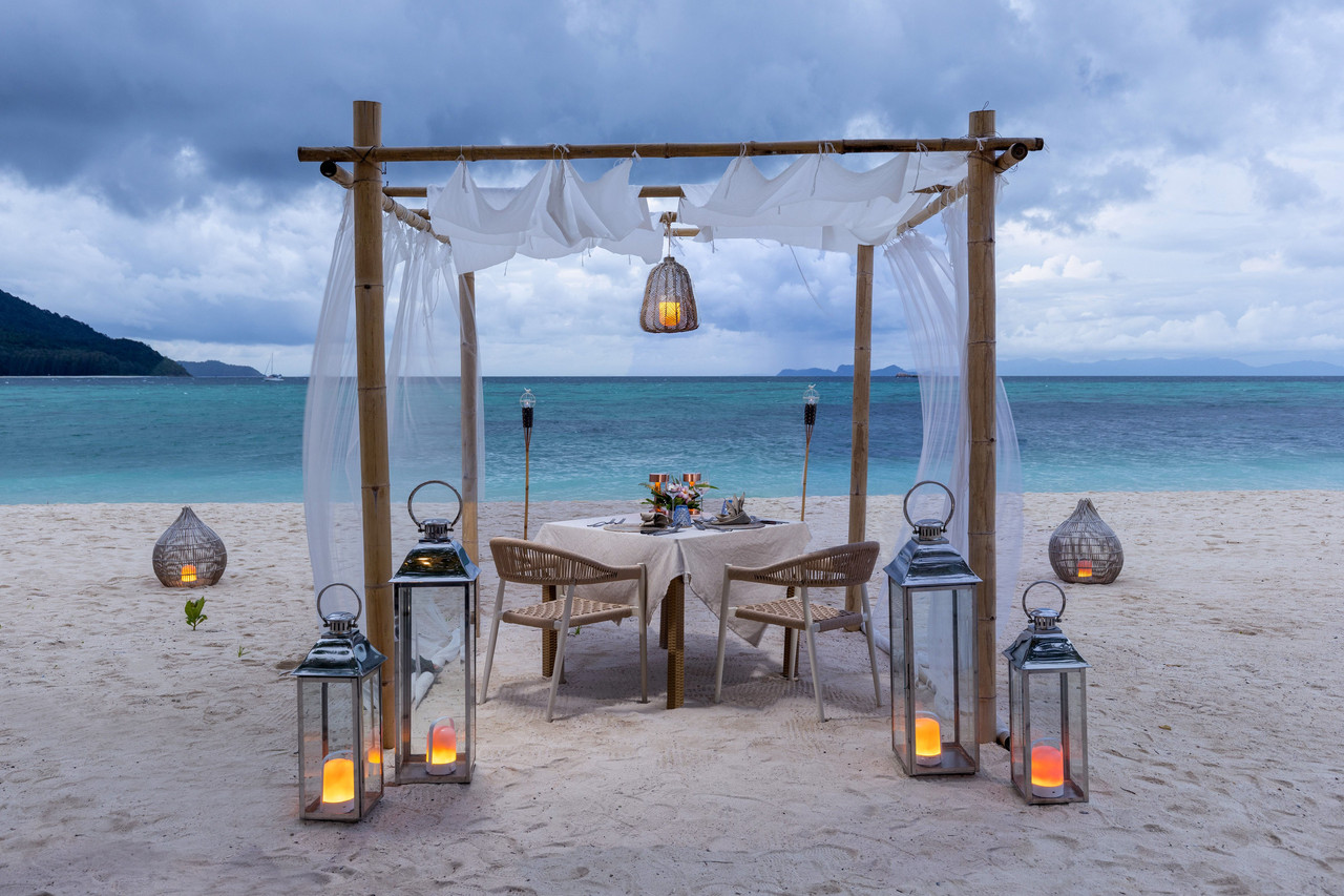 Private beachfront dining table on Sunrise Beach set with lanterns and white drapes facing the Andaman Sea